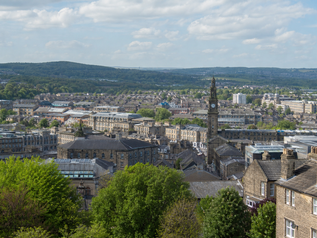 Aerial view of a town with a prominent clock tower, surrounded by green hills under a partly cloudy sky.