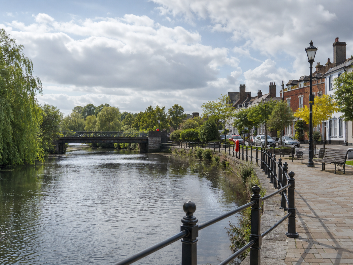 Riverside walkway with benches, trees, and classic houses, leading to a small bridge under a partly cloudy sky.